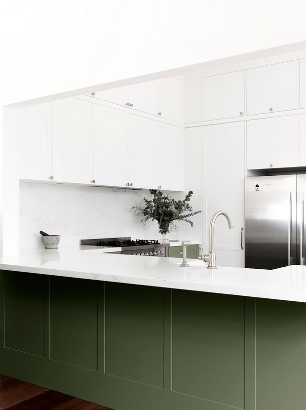 Bright, refined kitchen featuring extensive white shaker-style cabinetry, a large white marble-look island benchtop with an integrated sink and tall curved tap, pale veined splashback, stainless-steel refrigerator, gas cooktop, and a deep muted-green island base with panelled doors. Dark timber flooring adds contrast.