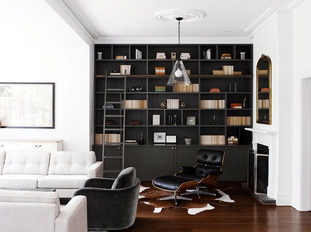 Refined living room with a large floor-to-ceiling dark built-in library and display shelving, ladder, white sofas, black lounge chair and ottoman, central pendant light, timber flooring, fireplace, and classic white architectural mouldings. The room has a high-contrast monochrome palette with warm wood accents and a sophisticated study-library feel.