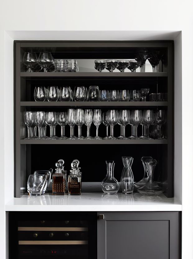 A built-in home bar cabinet with black open shelving displaying an extensive collection of wine glasses, tumblers, decanters, crystal bottles and spirits. The bar features a white stone countertop, dark cabinetry, and an integrated wine cooler below, presented in a refined, high-contrast monochrome palette.