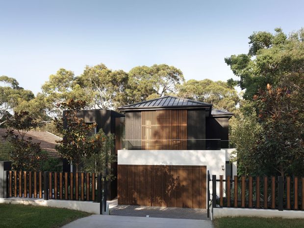 Street-facing view of a contemporary residential entrance and garage, set among mature trees. The dark facade combines vertical timber screening with charcoal cladding, a low-profile hipped metal roof, white rendered wall sections, and a timber-toned garage door. A slatted boundary fence and sliding gate frame the driveway.