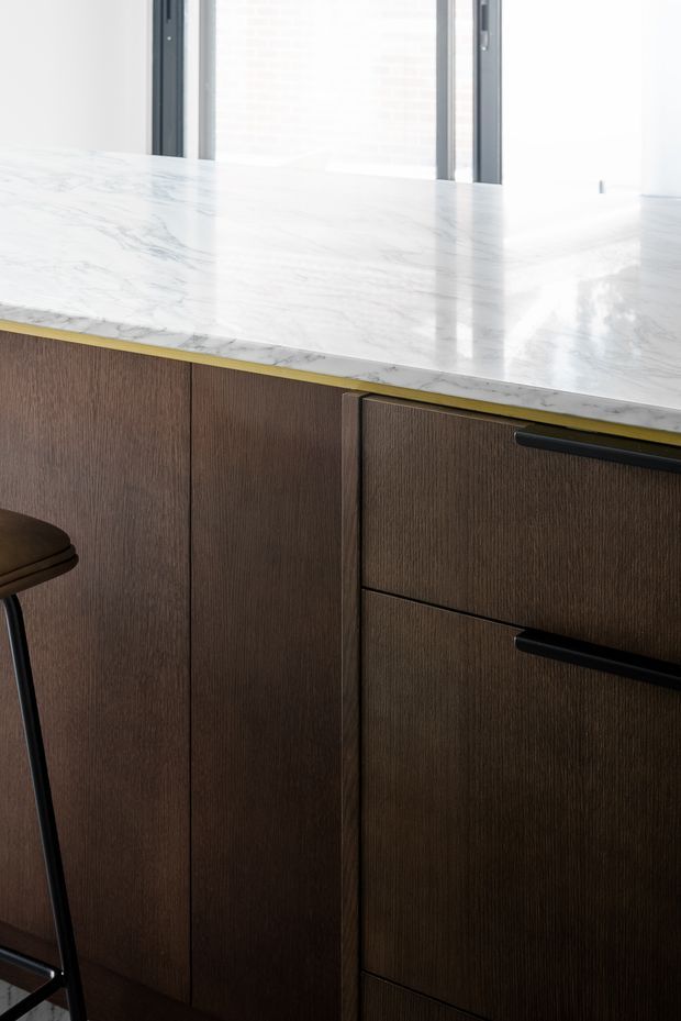 Close-up detail of a refined kitchen island or cabinet run, featuring dark vertically grained timber cabinetry, slim black bar handles, and a bright white marble benchtop with subtle grey veining. A partial dark bar stool is visible at the left, with narrow dark-framed glazing in the background.
