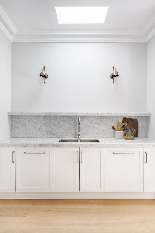 Bright, refined kitchen or butler’s pantry wall featuring white shaker-style cabinetry, a pale veined stone benchtop and full-height splashback, undermount sink with a slim metal tap, polished metal wall sconces, white walls with ornate crown moulding, a skylight, and light timber flooring.