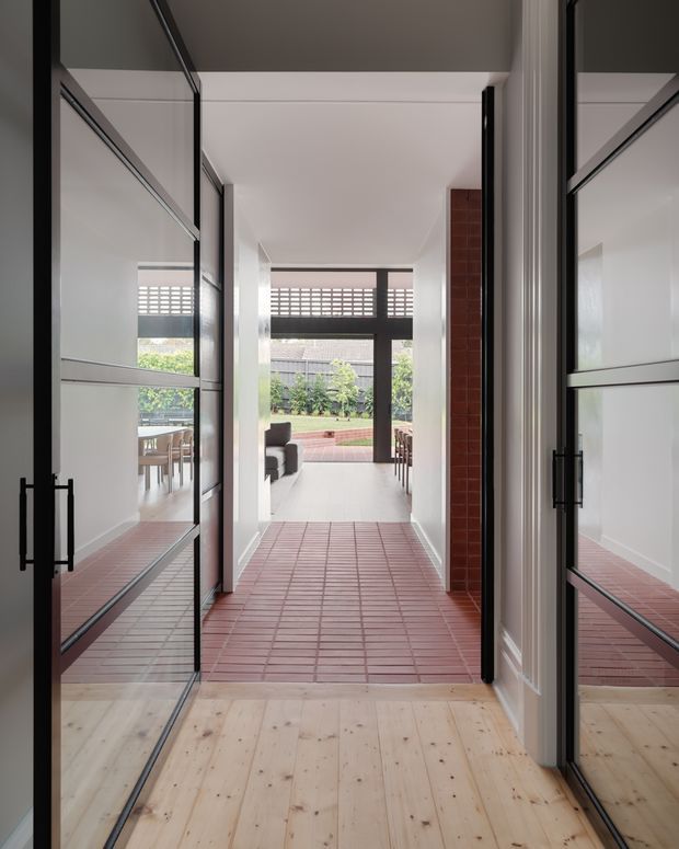 A bright residential hallway or entry passage viewed through a series of black-framed glazed doors. The foreground has pale timber flooring, transitioning to a red terracotta-toned tiled corridor. White walls and ceiling create a clean, modern interior, while large glazed openings reveal a dining area and living space leading to a landscaped outdoor courtyard. Reflections in the glazing emphasize the strong indoor-outdoor connection.
