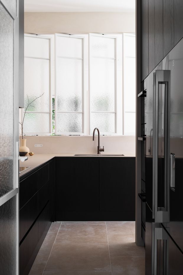 A compact, contemporary kitchen viewed through an open doorway. The room features matte black full-height cabinetry arranged in an L-shaped layout, a pale stone or solid-surface benchtop and splashback, a dark gooseneck mixer tap, and large white-framed textured-glass windows that provide bright diffused light. Warm taupe-grey large-format floor tiles contrast with the black joinery. A tall dark refrigerator or integrated appliance is visible on the right.