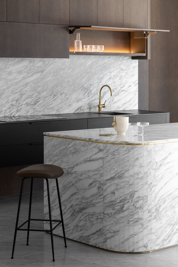 A sophisticated contemporary kitchen featuring a large marble-clad island with a rounded end, dark cabinetry, a veined marble splashback, and a slim brass edge detail. The island includes a raised seating area with a dark leather bar stool, while integrated cabinetry and an open illuminated display shelf provide storage and visual warmth. A brass gooseneck tap and minimal glassware add refined accents.