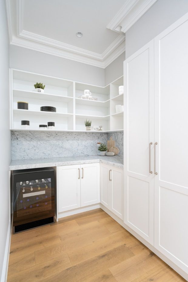 Bright, compact butler’s pantry with white built-in cabinetry, open display shelving, a grey veined stone benchtop and matching splashback, integrated wine fridge, timber flooring, and classic decorative ceiling cornices. The space has a refined, light neutral palette with brass-toned cabinet hardware and curated shelving decor.