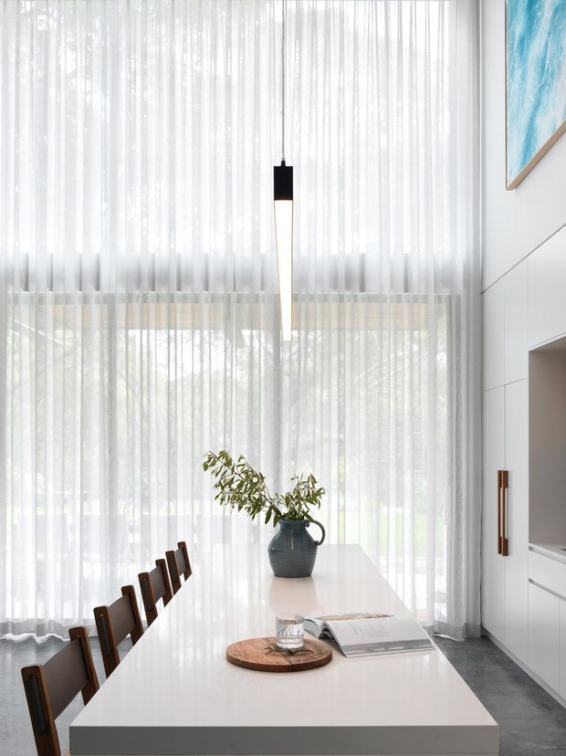 Bright, refined dining area centered on a long white rectangular dining table with dark timber dining chairs. Full-height sheer curtains create a softly diffused backdrop, while minimalist white built-in cabinetry and a slim linear pendant light reinforce the clean contemporary interior. A dark ceramic vase with branches, timber serving board, glass, and an open book provide restrained natural accents.