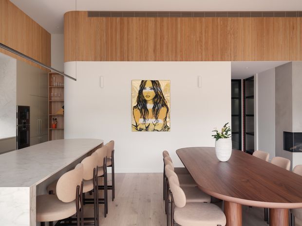 Contemporary open-plan kitchen and dining area with a large pale stone island, integrated cabinetry, timber floors, vertical timber wall panelling, and a sculptural rounded timber dining table. Soft upholstered dining chairs, understated pendant lighting, and a central artwork create a warm, refined interior. A fireplace is visible in the adjoining living area.