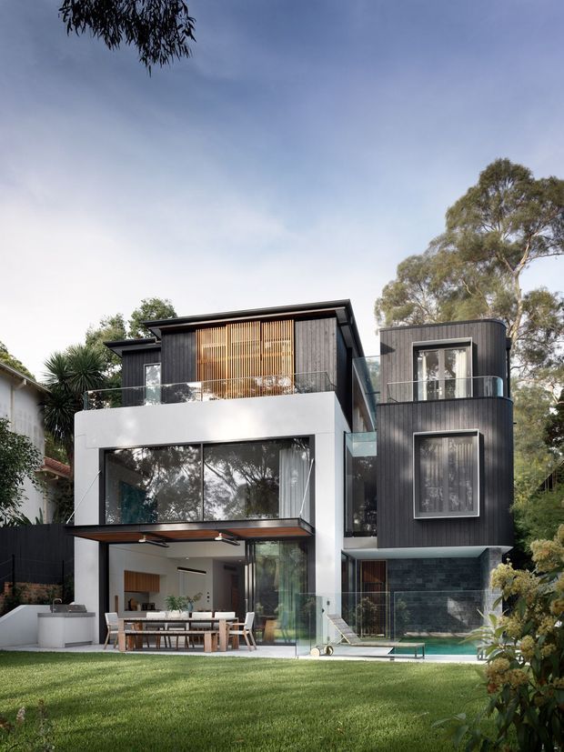 Exterior view of a contemporary multi-level residence set within a leafy suburban landscape. The house features dark vertical cladding, crisp white rendered sections, extensive glazing, projecting balconies with glass balustrades, timber screening, and a sheltered outdoor dining terrace beside a small swimming pool.