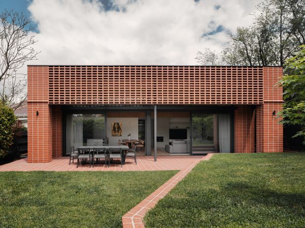 Exterior view of a compact contemporary brick pavilion or extension set in a landscaped garden. The facade features red brick walls, a large perforated brick lattice screen across the upper portion, and broad glazed sliding doors opening to a tiled terrace with an outdoor dining table. A brick path crosses the lawn toward the building, while the interior living area is visible through the glazing.