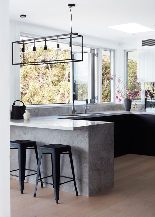 Bright contemporary kitchen interior with a large concrete-look island, black cabinetry, pale timber flooring, expansive windows, and a striking rectangular black metal pendant light. Two black bar stools sit at the island, while a sink and tap are positioned beneath the windows. The space is filled with natural light and overlooks leafy vegetation.
