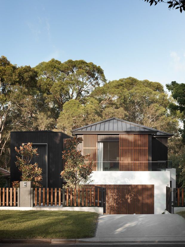 Front exterior view of a contemporary multi-level residence set among mature trees. The house combines dark vertical cladding, a pitched standing-seam metal roof, warm timber screening and fencing, a white rendered upper facade, glazed balcony balustrading, and an integrated garage. A concrete driveway and landscaped front lawn lead to the street.