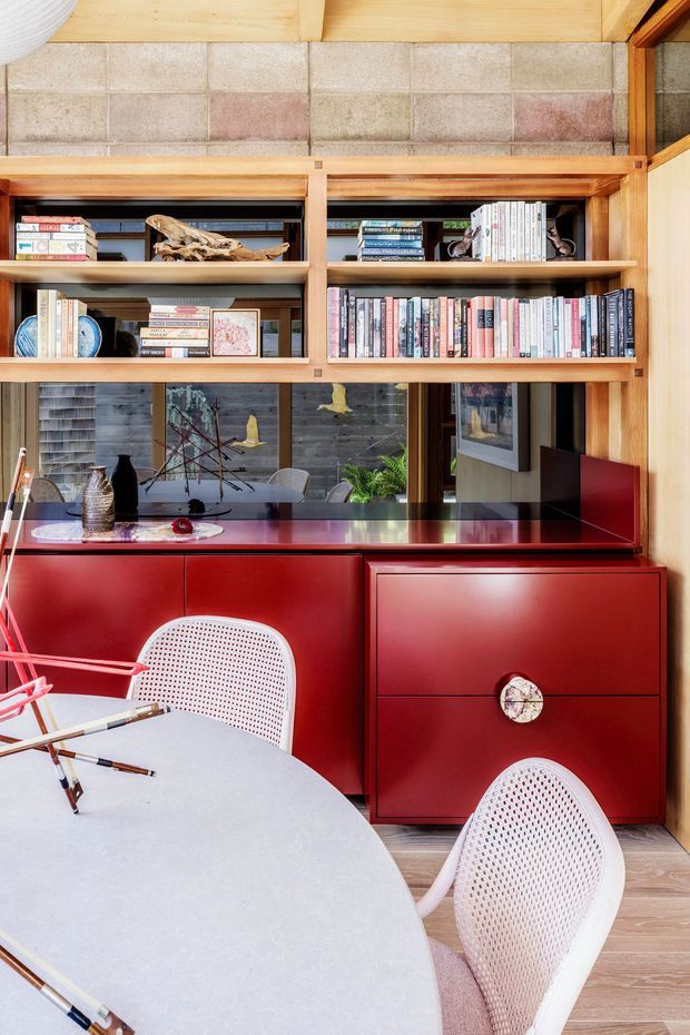 A compact, richly detailed dining and study area with a large round pale tabletop in the foreground, white woven chairs, and a striking glossy red built-in sideboard and cabinetry. Above the cabinetry is an open timber bookshelf filled with books and decorative objects. The room features warm timber framing, a tiled block-pattern wall, timber flooring, and extensive internal glazing or mirrored surfaces reflecting adjacent furnishings and greenery. The design combines strong mid-century influences with warm contemporary joinery and playful colour contrast.