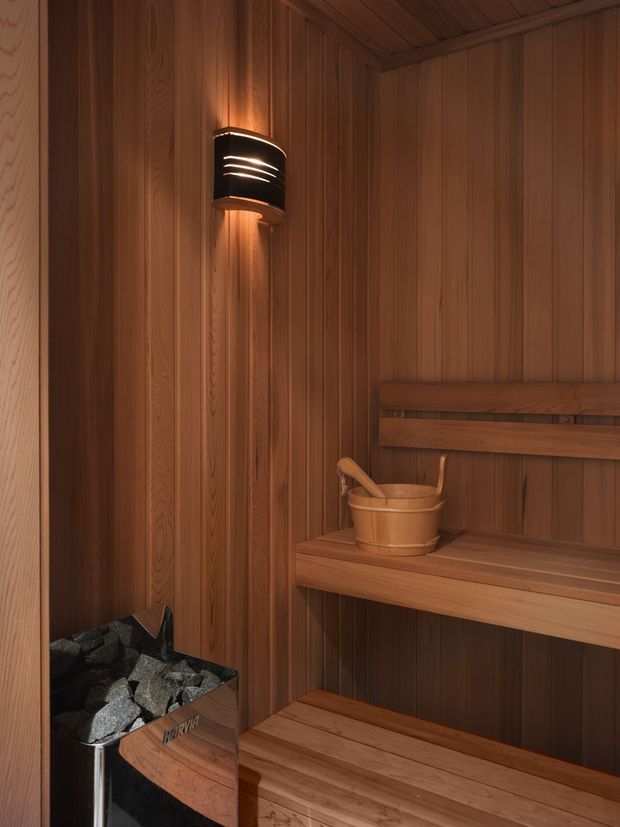 Compact wood-lined sauna interior with vertical timber wall and ceiling panelling, tiered timber benches, a stone-filled sauna heater, wooden bucket and ladle, and a warm wall-mounted light creating an amber glow.