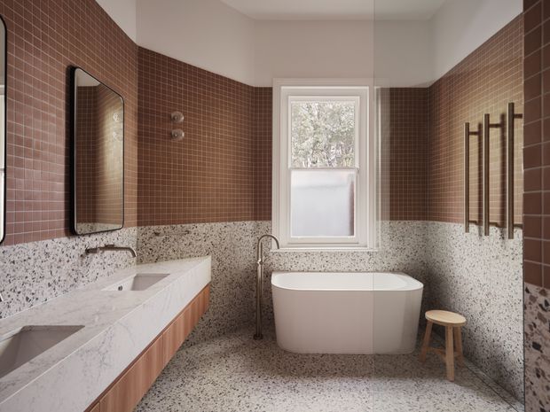 A refined bathroom with a freestanding white bathtub beneath a window, a double vanity with pale stone benchtops and timber cabinetry, warm brown small-format wall tiles, and terrazzo-style flooring and lower wall cladding. Dark heated towel rails and black-framed mirrors add contrast.