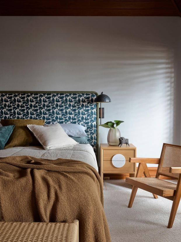 A warmly styled bedroom with a low upholstered bed, patterned blue-and-white feature headboard, layered white and brown bedding, and a mid-century timber bedside cabinet. A black dome wall lamp, leafy plant, sculptural ornament, woven timber lounge chair, and upholstered bench complete the room. Natural light filters across the pale wall, creating a calm, tactile interior.