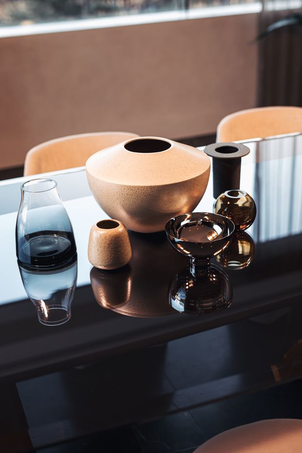 A close-up interior still life of sculptural ceramic and glass vessels arranged on a glossy black dining table. The setting includes curved upholstered dining chairs, a warm brown wall panel, and strong reflections across the tabletop. The composition has a refined, contemporary decorative aesthetic with warm neutral ceramics contrasted against dark reflective surfaces.