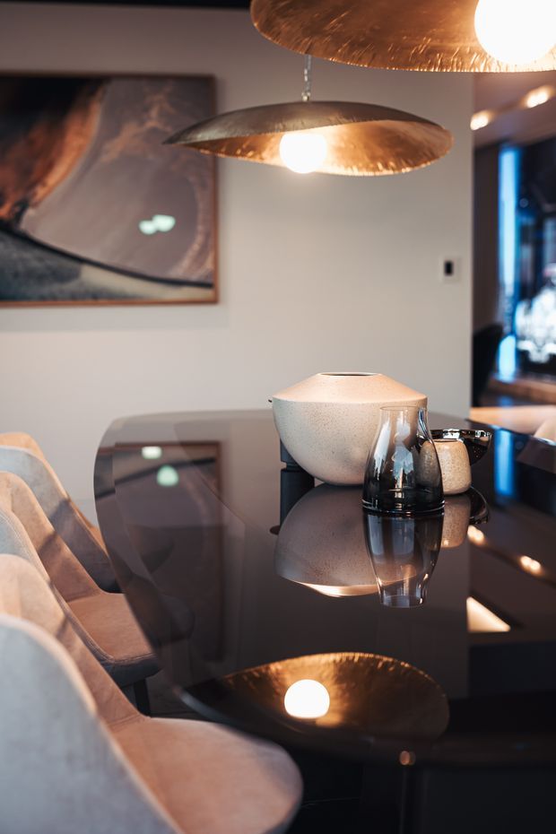 A warm, moody dining area centered on a glossy dark dining table with upholstered dining chairs, sculptural ceramic vessels, glassware, and distinctive copper-toned pendant lights. A large abstract artwork hangs on the wall, while a darker adjoining space is visible through an opening in the background. Reflections on the table create a dramatic, intimate atmosphere.