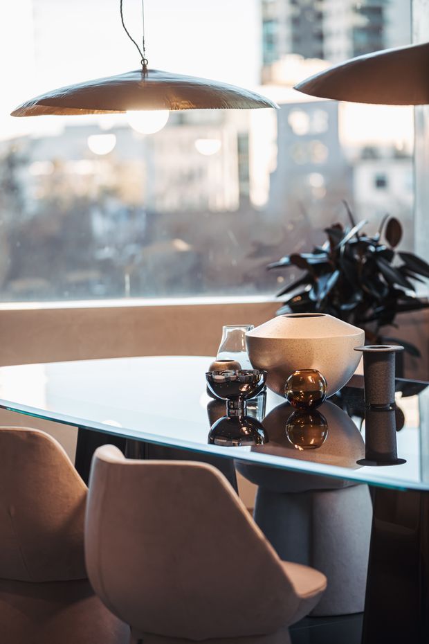A sophisticated dining area beside large glazing, featuring a glossy glass dining table with sculptural upholstered chairs, warm metallic and ceramic table decor, a large indoor plant, and oversized pendant lights. The blurred urban buildings beyond the window create an upscale city apartment atmosphere.