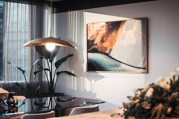 A warmly lit contemporary dining area with a glass-top table and upholstered chairs beside full-height sheer curtains. A sculptural floor lamp, large indoor plant, and abstract framed artwork create a refined, artistic atmosphere, with strong sunlight casting shadows across the pale wall.