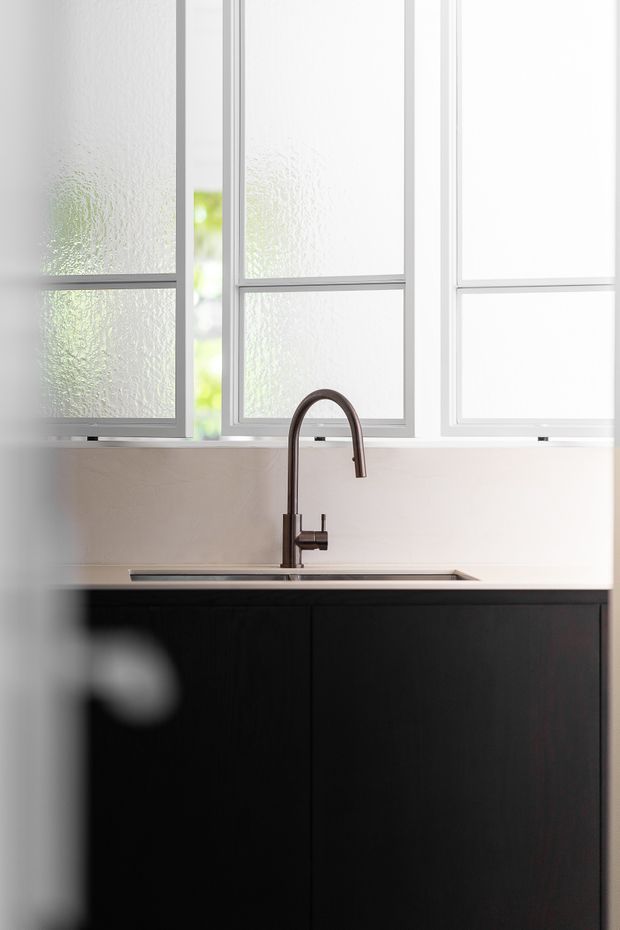 Close-up view of a minimalist kitchen sink area with a dark integrated cabinet run, pale smooth backsplash, tall arched dark metal mixer tap, and large white-framed textured-glass windows providing soft natural light.