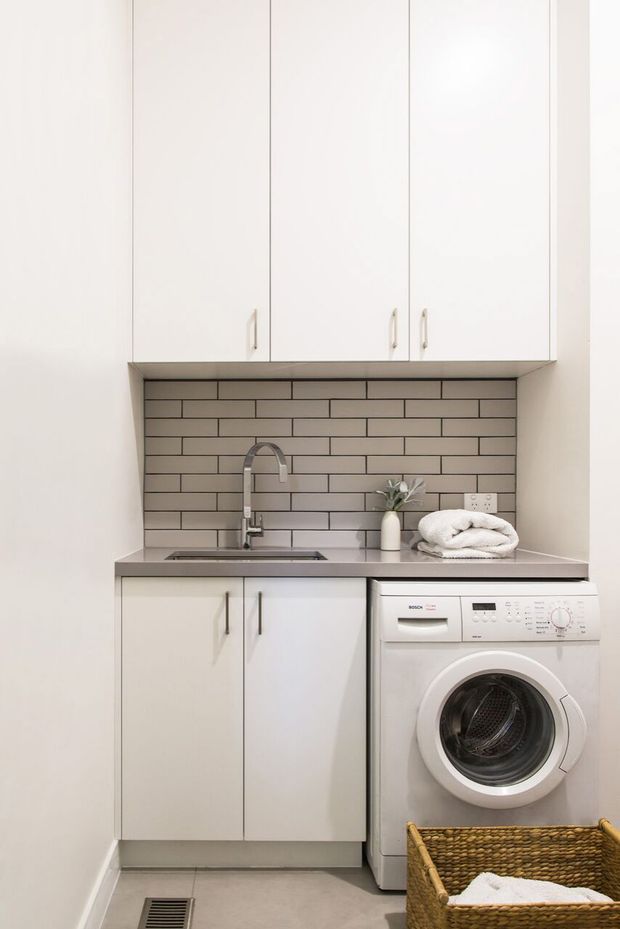 Compact, bright laundry room with white full-height overhead and lower cabinetry, a stainless-steel benchtop, grey subway-tile splashback, integrated sink and tap, front-loading washing machine, and woven laundry basket. The space has a clean, practical contemporary fit-out with neutral tones.