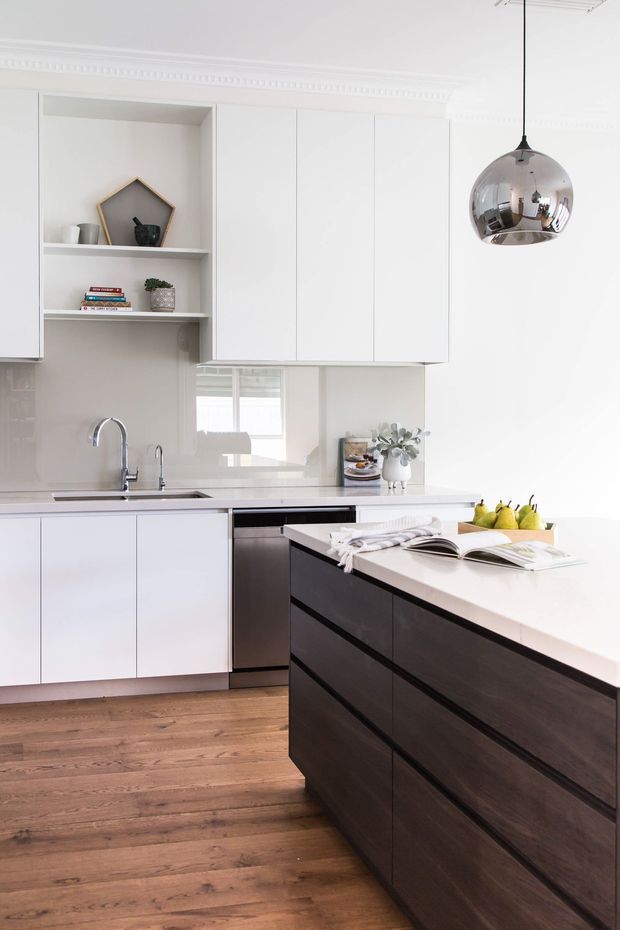Bright contemporary kitchen with white handleless cabinetry, dark timber island joinery, a pale stone benchtop, glossy light backsplash, timber flooring, and a reflective chrome pendant light. Decorative open shelving and traditional ceiling cornice add subtle classic detailing.
