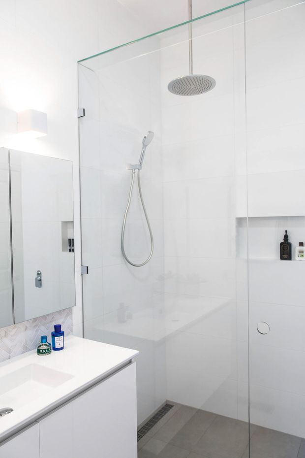 Bright, minimalist bathroom featuring a large frameless glass walk-in shower with overhead rain shower and handheld fixture, white tiled walls, a recessed shelf, grey tiled floor, and a white floating vanity with integrated basin and mirror.