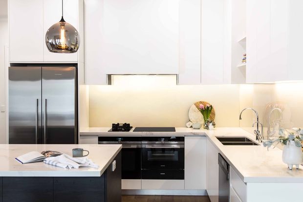 Bright contemporary white kitchen with a U-shaped layout and central island. The space features sleek white cabinetry, dark lower cabinetry, stainless-steel double refrigerator, integrated black oven and cooktop, pale stone-look benchtops, and a softly illuminated splashback. A reflective pendant light hangs above the island, with minimal styling and open shelving.