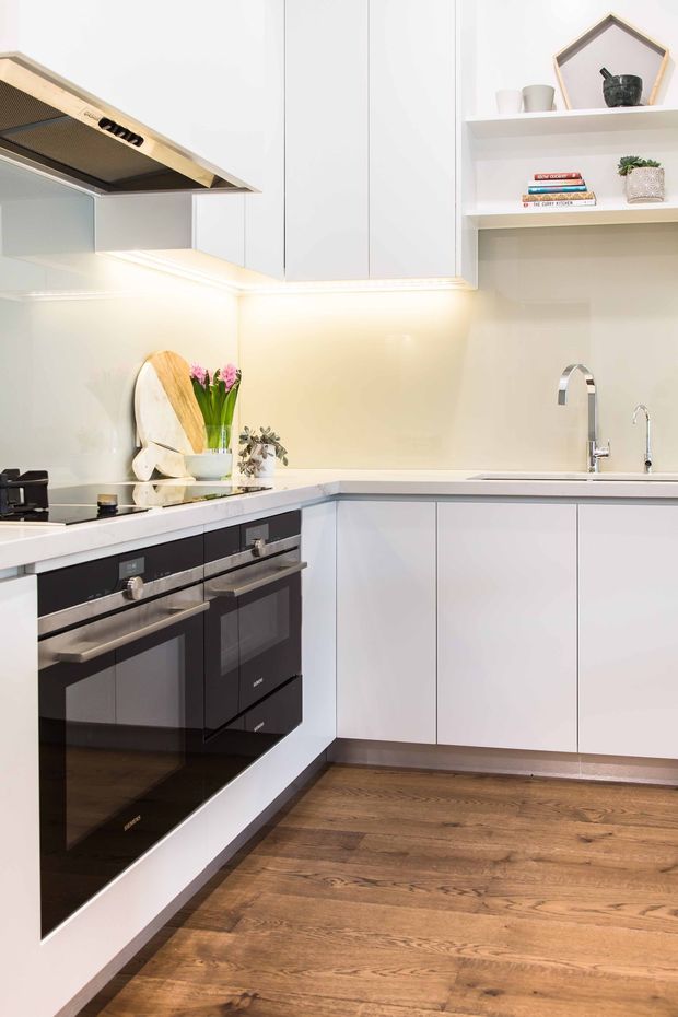 Bright contemporary L-shaped kitchen with white flat-panel cabinetry, integrated under-cabinet lighting, pale benchtops and backsplash, black built-in cooking appliances, stainless-steel tapware, open display shelving, and warm timber flooring. Minimal decorative styling includes cutting boards, flowers, books, and small objects.