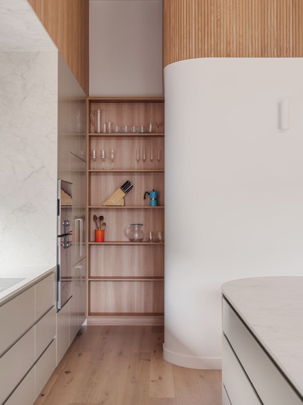 A refined contemporary kitchen featuring pale marble-look benchtops and splashback, warm timber cabinetry and open shelving, integrated appliances, and a distinctive curved white partition or cabinetry element. Glassware and kitchen accessories are displayed on the timber shelves, while light timber flooring and vertical timber slats add warmth and texture.