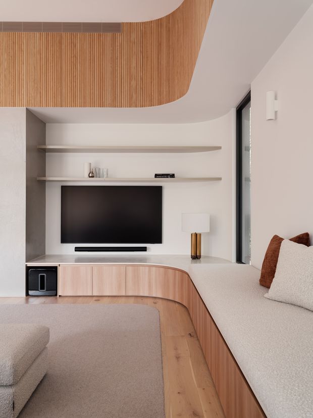 A refined contemporary living room with a built-in curved timber entertainment unit and integrated upholstered window seat. The room features pale textured walls, warm light-wood cabinetry and ceiling slats, open display shelves, a wall-mounted television, soft neutral upholstery, and dark-framed floor-to-ceiling glazing. Rounded joinery and ceiling details create a seamless, sculptural interior.