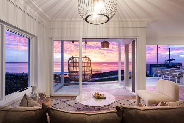 A bright, refined coastal living room with panoramic glazing overlooking the ocean at sunset. The room features a large sectional sofa, two woven hanging chairs, a round marble-topped coffee table, patterned rug, sculptural pendant lighting, and white painted walls with decorative trim. An adjoining outdoor terrace with dining furniture is visible through the open glazing.