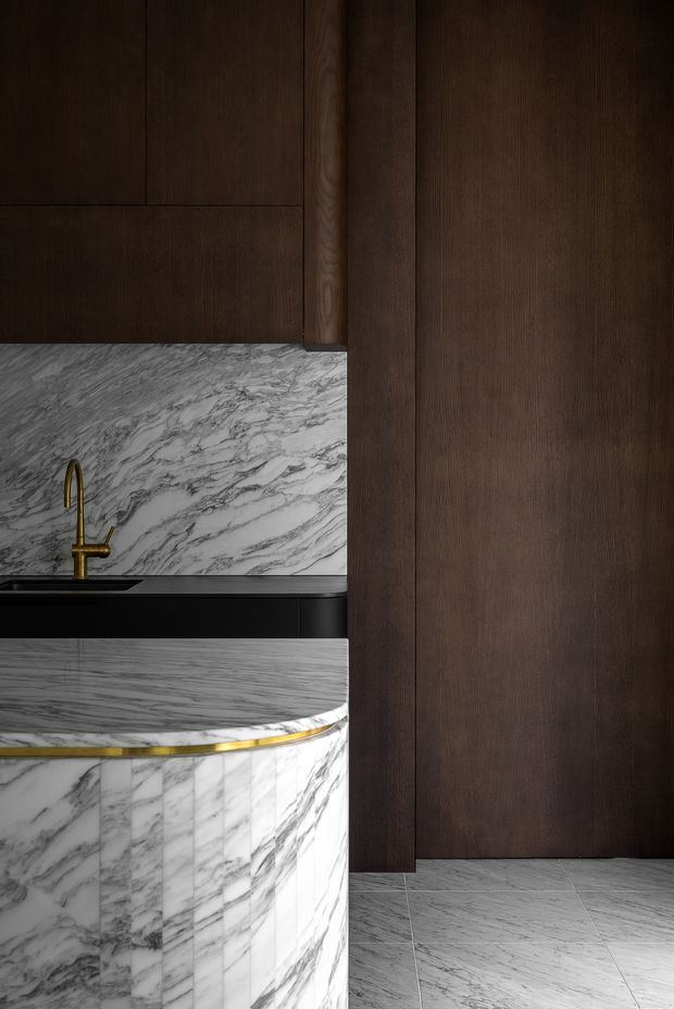 Close-up view of a refined contemporary kitchen featuring dark timber cabinetry, a dramatic white-and-grey veined marble splashback and curved marble island, with a slim brass trim and matching brass gooseneck tap. The composition is dark, luxurious and highly material-focused.