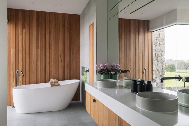A refined contemporary bathroom featuring a freestanding white bathtub, double vessel basins on a long timber vanity, matte black tapware, grey floor tiles, and extensive vertical timber wall panelling. A large mirror reflects the vanity and bath, while a stone-clad exterior wall and expansive glazing provide a connection to the landscape.