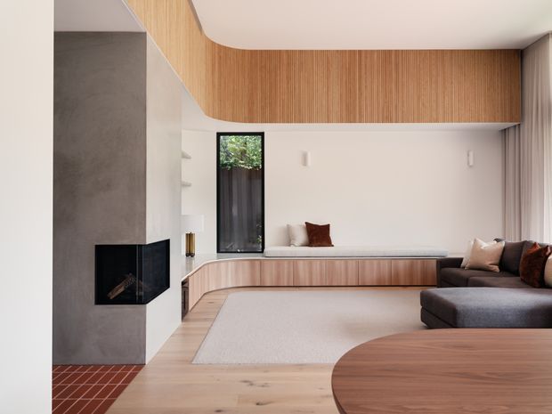 A refined contemporary living room with a sculptural built-in timber feature wrapping across the ceiling and rear wall, integrated window seating, and curved timber cabinetry. The room includes a minimalist black-framed fireplace set into a textured grey wall, a large neutral rug, a dark sectional sofa, and a rounded timber coffee table. The palette is warm and neutral, with strong emphasis on natural timber and curved built-in forms.