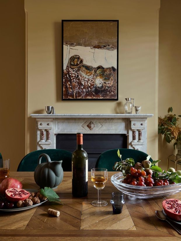 A warm, traditionally styled dining room centered on a richly patterned timber dining table set with wine, fruit, glassware, and ceramic vessels. A carved white fireplace mantel with marble-like surfaces anchors the room, with deep green upholstered dining chairs and a framed abstract artwork above. The palette is earthy and moody, with ochre walls, dark wood, burgundy fruit tones, and antique-inspired details.