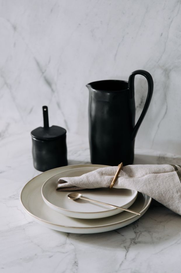 Still-life product photograph of a minimalist table setting on a pale white-grey veined marble surface. Stacked neutral ceramic plates, a folded natural linen napkin with a wooden napkin ring, a metal spoon, and matte black ceramic serving vessels are arranged against a matching marble backdrop.