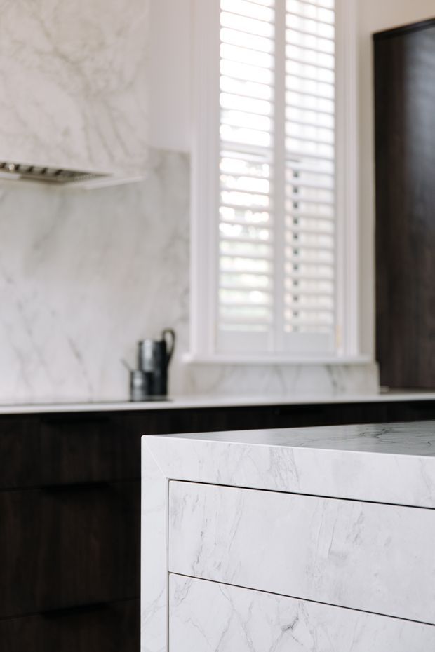 Close-up view of a refined kitchen featuring a large white marble-clad island with integrated drawer fronts, dark timber cabinetry, a white marble backsplash, and a bright window fitted with white plantation shutters. A dark rangehood and small countertop appliance are visible in the background.