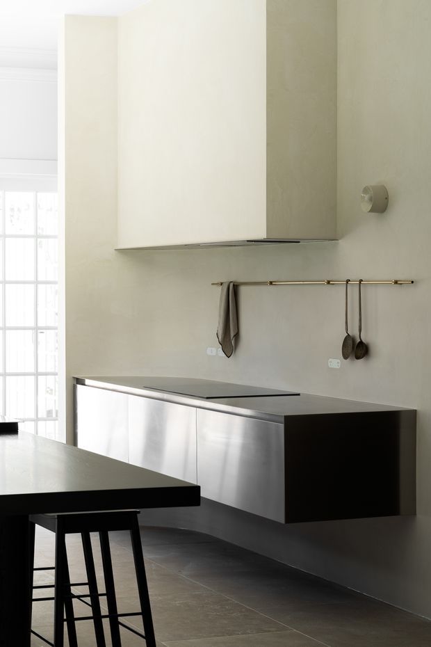 A minimalist contemporary kitchen with a floating stainless-steel base cabinet and integrated dark benchtop, a large concealed rangehood enclosure, and a slim brass rail for hanging utensils and a tea towel. The space has pale plaster walls, large dark floor tiles, a black island or peninsula with a simple black stool, and abundant natural light from tall multi-pane glazing.