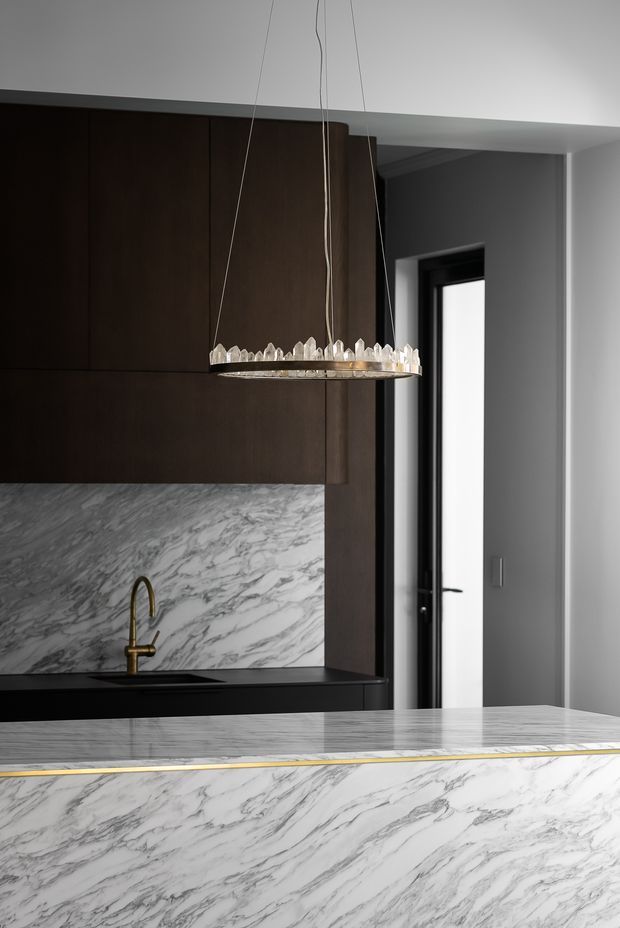 A refined contemporary kitchen featuring dark timber cabinetry, dramatic white-and-grey veined stone splashback and waterfall island, a black benchtop, dark-framed internal glazing, and a sculptural crystal pendant light. Brass tapware provides a subtle accent against the monochromatic material palette.