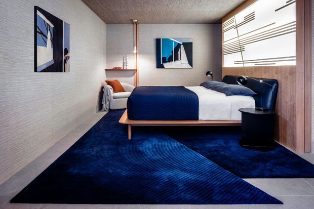 A compact, sophisticated bedroom with a low-profile timber platform bed, navy upholstered headboard, white bedding, and deep navy carpeting. Textured light walls, a warm timber ceiling, abstract artwork, a small black bedside table, and a sculptural wall-mounted bedside light create a refined contemporary interior. A partially screened or slatted timber wall feature is visible behind the bed.