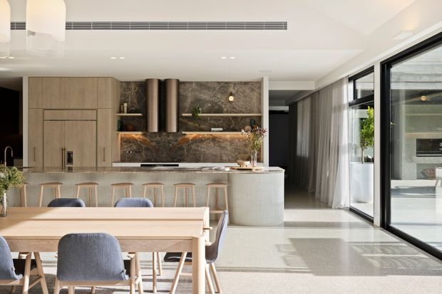 A bright, open-plan contemporary interior combining a kitchen, dining area and living zone. The kitchen features tall light timber cabinetry, a large curved island with a pale ribbed finish, a dark veined stone backsplash and benchtop, and metallic cylindrical rangehoods. A timber dining table with upholstered chairs occupies the foreground, while full-height black-framed glazing opens to an exterior terrace or courtyard. Polished light flooring, neutral finishes and abundant daylight create a refined, airy atmosphere.