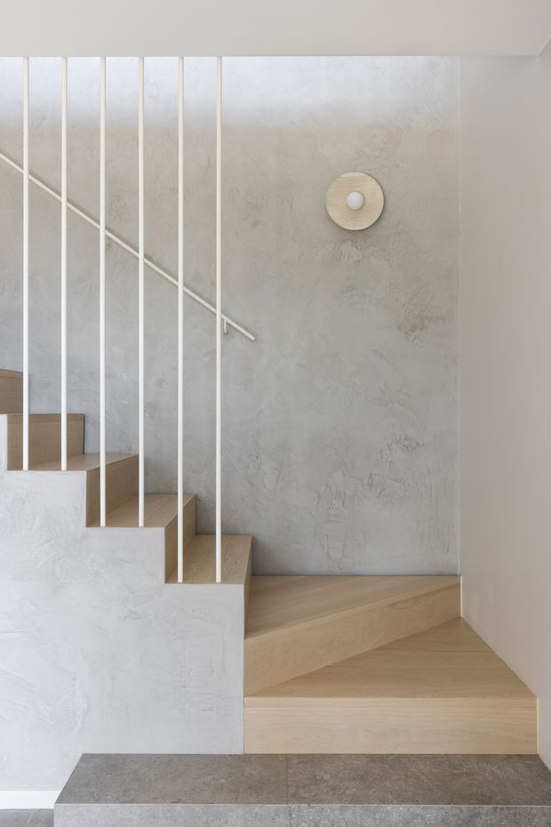 A minimalist interior stairway with pale timber stair treads, a textured light-grey plaster wall, and slender white vertical balusters with a simple diagonal handrail. A round wall light adds a subtle feature, while dark grey stone or concrete flooring anchors the base of the stairs.