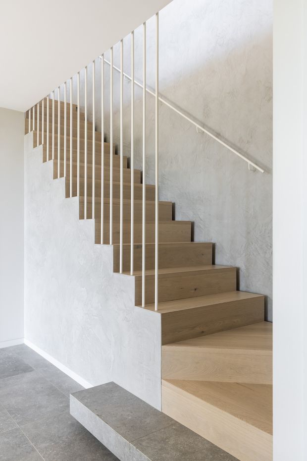 Bright minimalist stairway with warm light-wood treads and risers, a textured white plaster wall, and slender white metal vertical balusters. The staircase turns along the wall and sits above a grey stone-tile floor with a raised platform at its base.