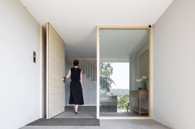 A bright, minimalist residential entryway with a large pivot timber door opening toward a stair hall. The space features pale walls, grey tiled flooring, a glass-framed internal partition, a timber console, and views through glazing toward a leafy outdoor setting. A staircase rises beyond the entry, with a person walking through the space.