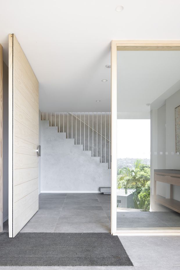 Bright contemporary interior entry with a large timber pivot door, grey tiled floor, concrete stair, and slim vertical metal balustrade. A full-height glazed opening reveals a small landing or study area with timber joinery and greenery visible outside.