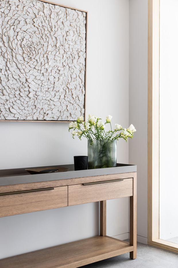 A bright, refined entryway vignette featuring a slim timber console with drawers, a dark stone-like top, open lower shelf, and slender metal handles. A textured monochromatic artwork hangs above, complemented by a ribbed glass vase with white flowers and a small dark decorative vessel. A large timber-framed glazed opening is visible at right.