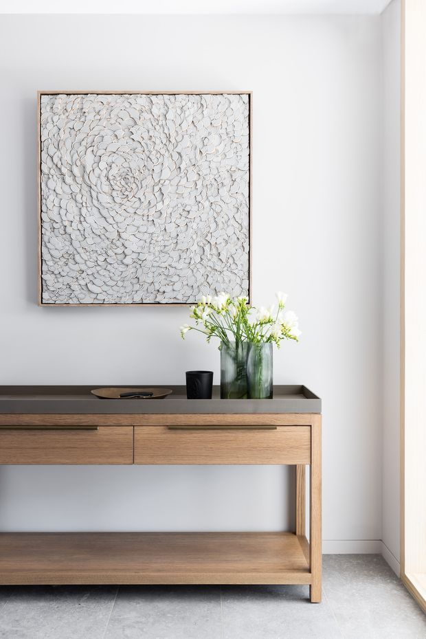 Bright, minimalist entryway featuring a slim walnut console with drawers and an open lower shelf, topped with a dark stone surface. A large textured monochromatic artwork hangs above, accompanied by glass vases and simple floral arrangements. Pale walls, light stone flooring, and strong natural light create a clean, refined atmosphere.