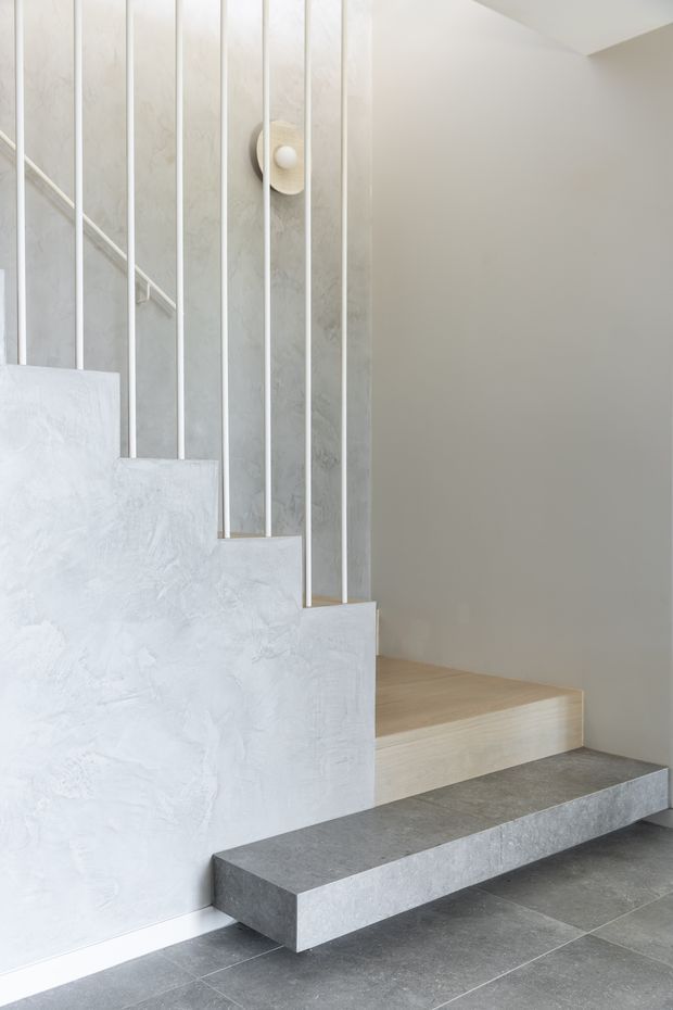 Minimalist interior stairway with pale textured plaster walls, slim white vertical balustrade rods, light timber stair treads, and a projecting dark grey stone or concrete step above large grey floor tiles. A simple wall light adds a subtle decorative detail.